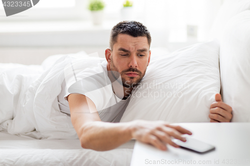 Image of young man reaching for smartphone in bed