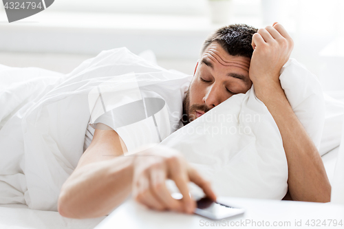 Image of young man reaching for smartphone in bed