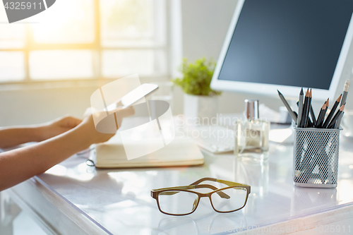 Image of Close-up of female hands using smart phone while working on computer at modern office interior