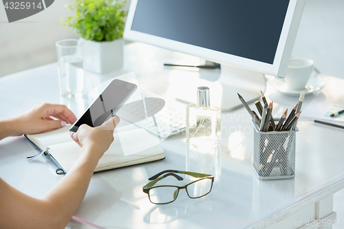 Image of Close-up of female hands using smart phone while working on computer at modern office interior