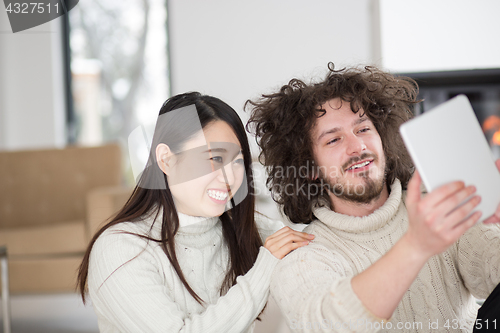 Image of multiethnic couple using tablet computer in front of fireplace