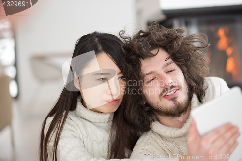 Image of multiethnic couple using tablet computer in front of fireplace