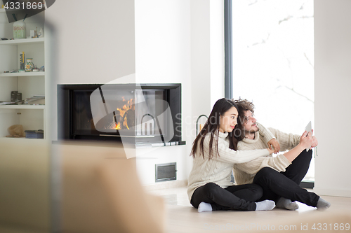 Image of multiethnic couple using tablet computer in front of fireplace