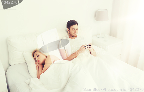 Image of man texting message while woman is sleeping in bed