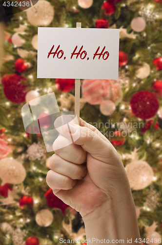 Image of Hand Holding Ho Ho Ho Card In Front of Decorated Christmas Tree.