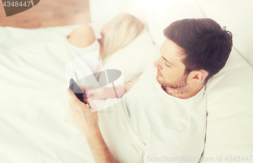 Image of man texting message while woman is sleeping in bed