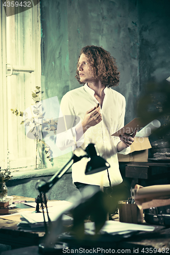 Image of Writer at work. Handsome young writer standing near the table and making up something in his mind