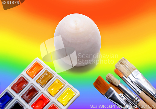 Image of Wooden not painted egg on the background of a rainbow and a brush with paints, the concept of coloring the world with paints