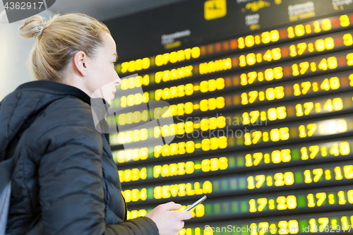 Image of Woman at airport in front of flight information board checking her phone.