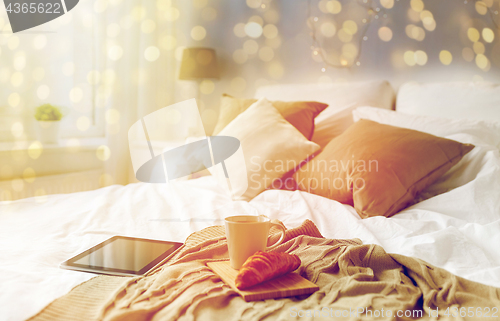 Image of tablet pc, coffee cup and croissant on bed at home