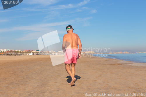 Image of Shirless senior man in shorts walking on sandy beach.