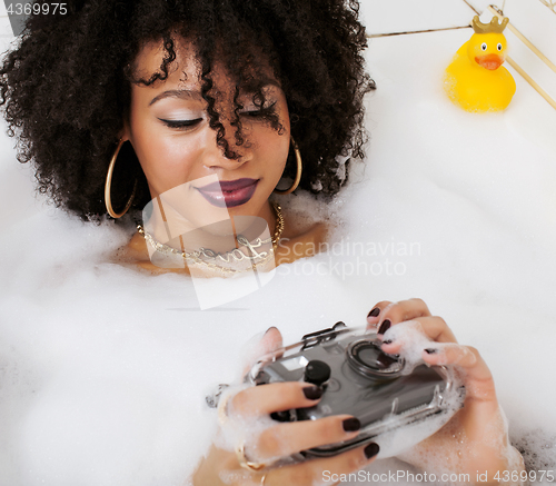 Image of young afro-american teen girl laying in bath with foam, wearing swag jewelry flawless, making selfie, modern lifestyle concept