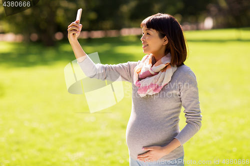 Image of happy pregnant asian woman taking selfie at park