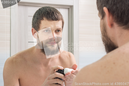 Image of An unshaven man applies a shaving foam on the palm of his hand, and looks in the mirror in the bathroom