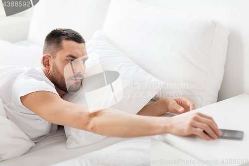 Image of young man reaching for smartphone in bed