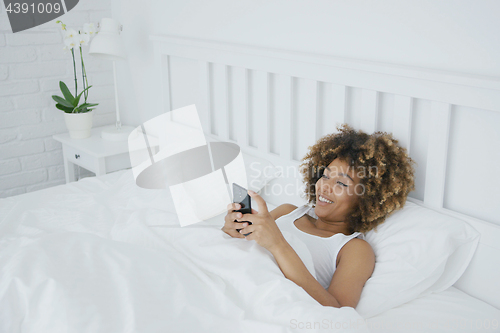 Image of Smiling woman relaxing with phone in bed