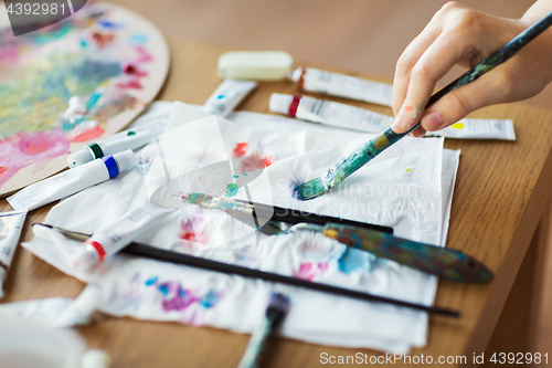 Image of artist hand with paintbrush, paper and paint tubes