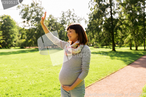 Image of happy pregnant asian woman waving hand at park