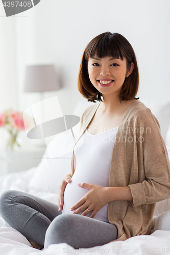 Image of happy pregnant asian woman sitting in bed at home