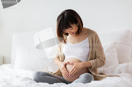 Image of happy pregnant woman making heart gesture in bed