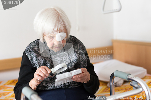 Image of Visually impaired elderly 95 years old woman sitting at the bad trying to read with magnifying glass.