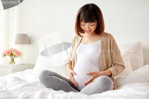 Image of happy pregnant asian woman sitting in bed at home