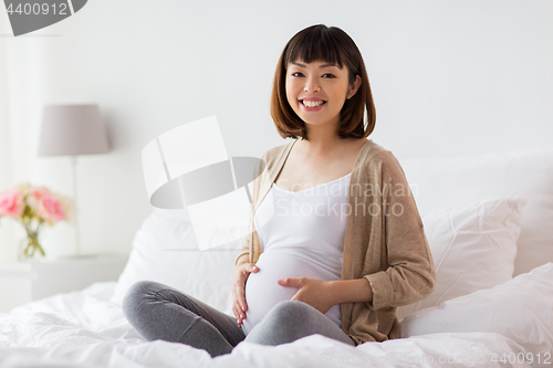 Image of happy pregnant asian woman sitting in bed at home