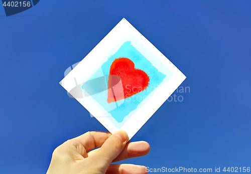 Image of Hand holding a picture of a heart against the blue sky