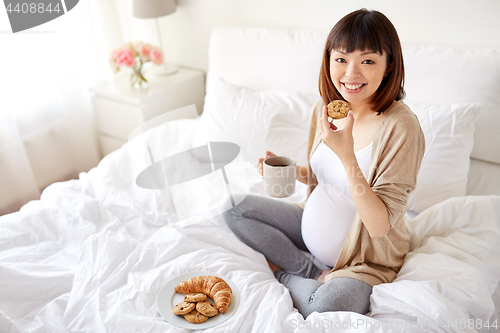 Image of happy pregnant woman eating cookie in bed at home