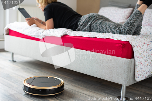 Image of Vacuum cleaning robot with relaxed woman reading in the background