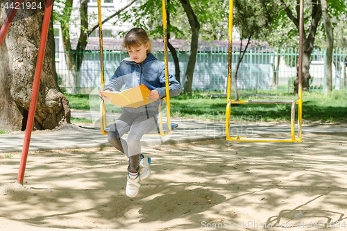 Image of A girl on a walk reading a book swinging on a swing at the playground