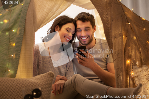 Image of happy couple with smartphone in kids tent at home