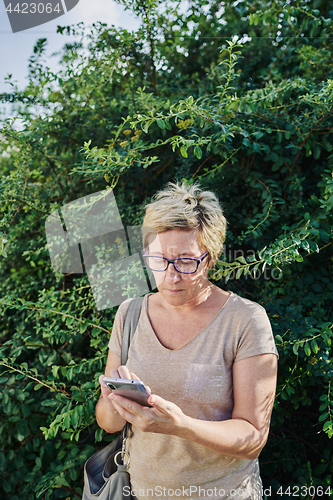 Image of Senior woman using smartphone near bush