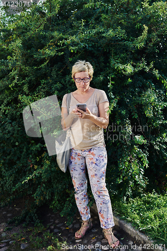 Image of Senior woman using smartphone near bush