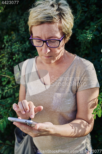 Image of Senior woman using smartphone near bush