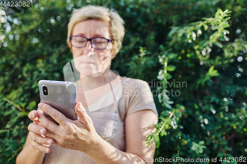 Image of Senior woman using smartphone near bush