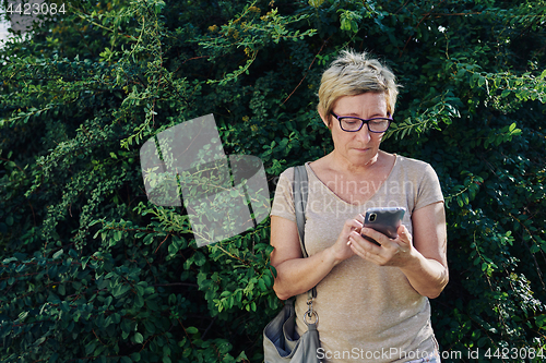 Image of Senior woman using smartphone near bush