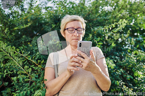 Image of Senior woman using smartphone near bush