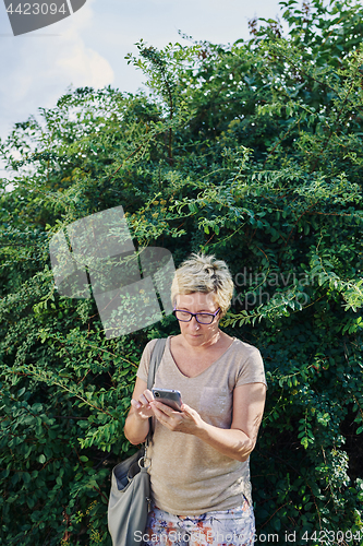 Image of Senior woman using smartphone near bush