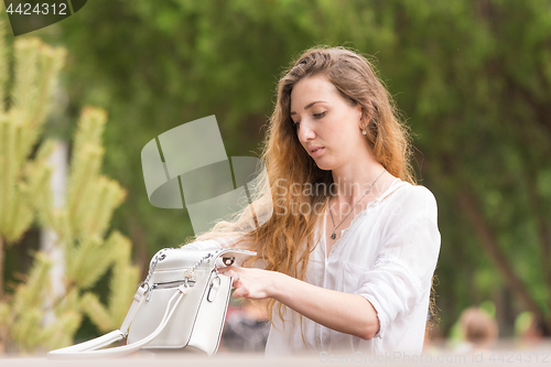 Image of Beautiful young girl is delving into her purse in the park