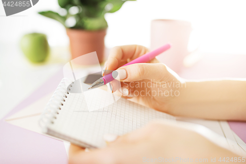 Image of The female hands holding pen. The phone on trendy pink desk.