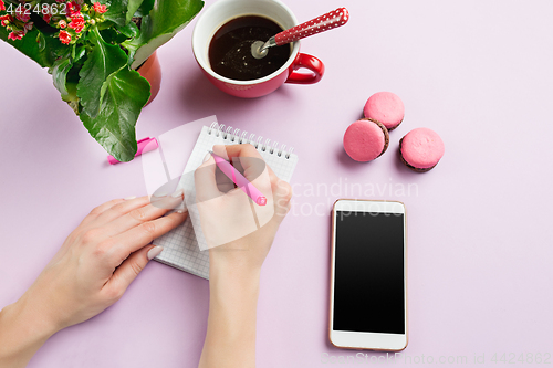 Image of The female hands holding pen. The phone and french macarons on trendy pink desk.