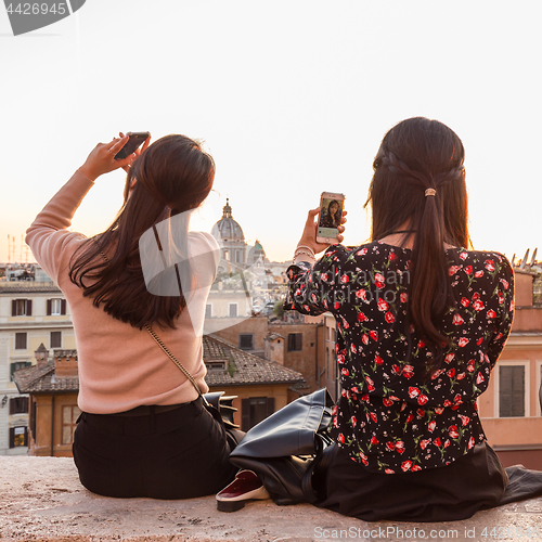 Image of Female tourists taking mobile phone photos of Piazza di Spagna, landmark square with Spanish steps in Rome, Italy at sunset.