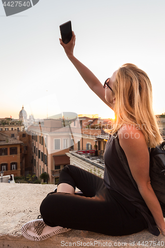 Image of Female tourist taking mobile phone photo of Piazza di Spagna, landmark square with Spanish steps in Rome, Italy at sunset.