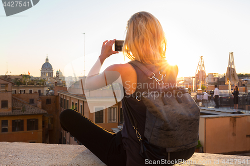 Image of Female tourist taking mobile phone photo of Piazza di Spagna, landmark square with Spanish steps in Rome, Italy at sunset.