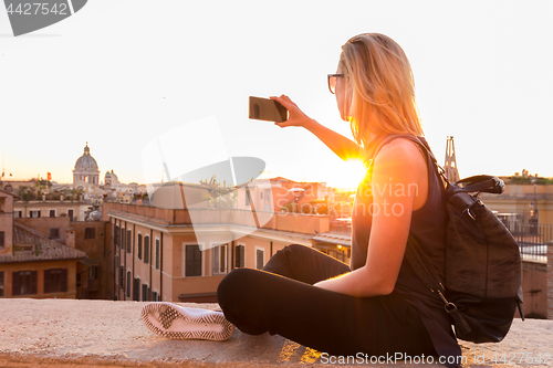 Image of Female tourist taking mobile phone photo of Piazza di Spagna, landmark square with Spanish steps in Rome, Italy at sunset.