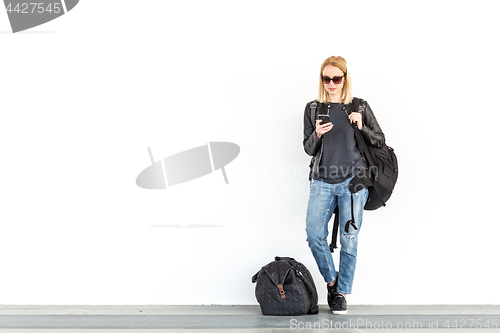 Image of Fashionable young woman using her mobile phone while standing and waiting against plain white wall on the station whit travel bag by her side.