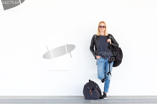 Image of Fashionable young woman standing and waiting against plain white wall on the station whit travel bag by her side.