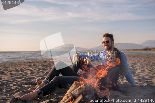 Image of Young Couple Sitting On The Beach beside Campfire drinking beer