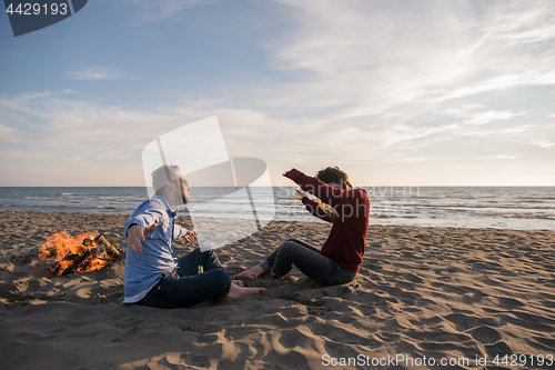 Image of Young Couple Sitting On The Beach beside Campfire drinking beer